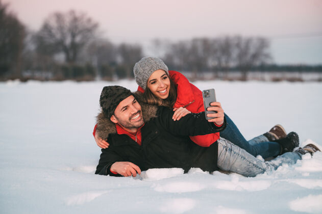 寒冷一对穿着暖和衣服的快乐的年轻夫妇一起躺在雪地上 一边用智能手机自拍 一边在冬天的乡间玩耍捕捉喜欢债券