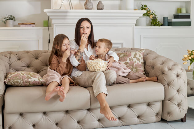 电影妈妈和儿子女儿坐在沙发上看电影一个女人 一个男孩和一个女孩一边看电影一边吃爆米花这家人周末在家休息女儿女人父母