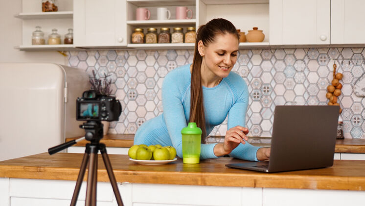 技术厨房里拿着笔记本电脑的年轻女子微笑着美食博客的概念一位女士正在录制一段关于健康饮食的视频三脚架上的相机媒体美丽家庭主妇