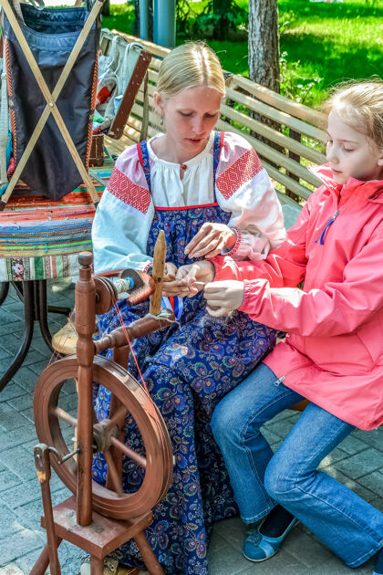 编织一个穿着民族服装的女人教一个女孩在手纺车上工作车轮木材自然