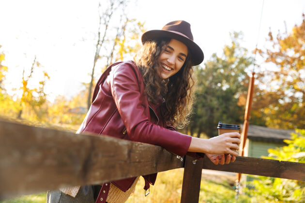 温暖美丽的年轻女子戴着帽子 斜靠在秋日公园的木桥上 端着一杯外卖咖啡美丽微笑女人