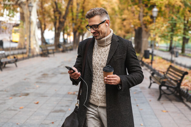 听力帅哥穿着外套走在户外 用耳机和手机听音乐 拿着外卖咖啡杯时尚咖啡休闲