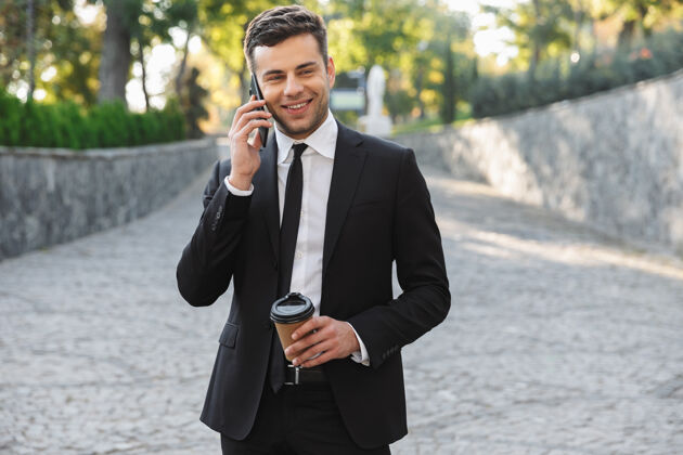 西装一位英俊快乐的年轻商人在商务中心附近的户外散步 一边用手机聊天一边喝咖啡高管领导公司