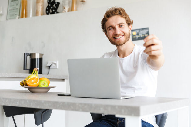 休闲快乐的小胡子男人在家里的桌子旁用手提电脑拿着信用卡聊天信贷吸引笔记本电脑