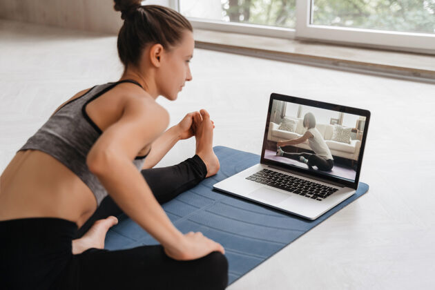远程适合穿着运动服的女士在家里用笔记本电脑在运动垫上做瑜伽教程.女教练在电脑上有一个虚拟瑜伽课女人体操私人教练