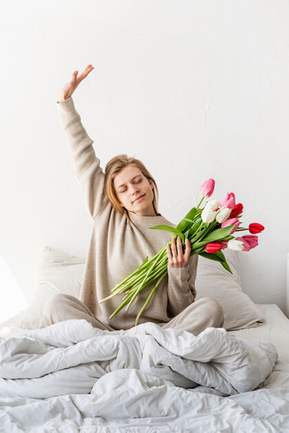 春天快乐的女人穿着睡衣坐在床上 愉快地享受鲜花和伸展白天惊喜室内