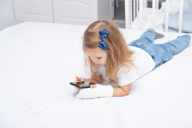 石膏手拿石膏的小女孩躺在床上用智能手机 看动画片或教育视频医学女孩治疗