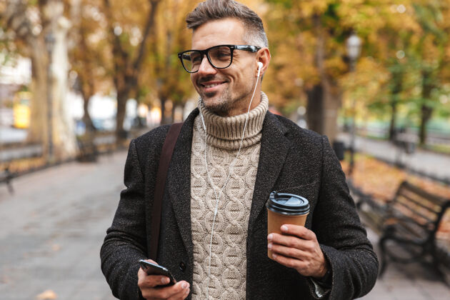 户外帅哥穿着外套走在户外 用耳机和手机听音乐 拿着外卖咖啡杯耳机听力外套