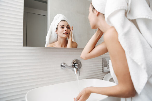 吸引头上戴着毛巾的漂亮年轻女子站在浴室里 对着镜子检查自己的脸手指不安治疗
