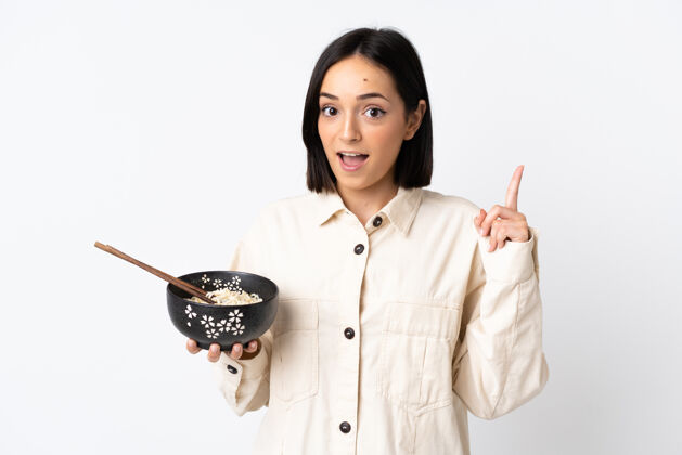 看一位年轻的白人妇女站在白色的墙上 一边用筷子端着一碗面条 一边指着一个好主意女人点人