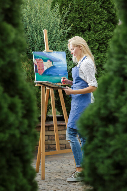 幻想美丽的金发女画家手里拿着画笔在花园里的画布上画画女孩艺术家自然