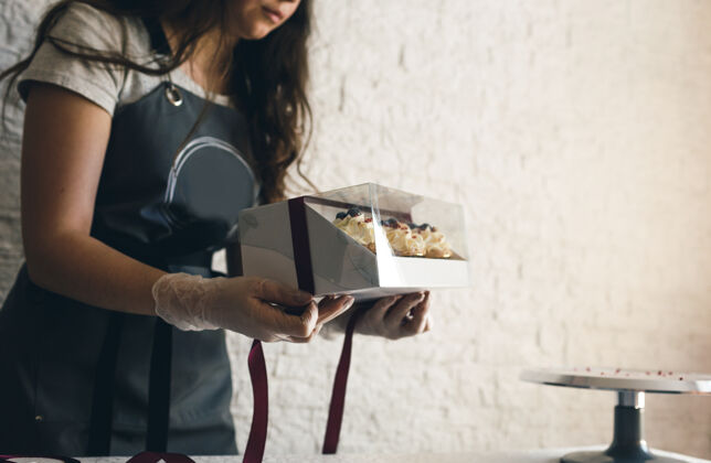 回收一个穿着灰色围裙的女孩在一个礼品盒里包上奶油纸杯蛋糕 把订单送到餐厅客户.烘焙在家里小吃聚会制服