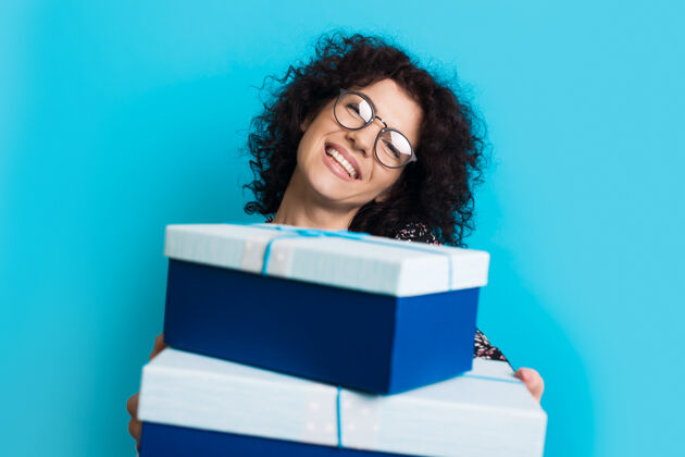 盒子一位留着卷发 戴着眼镜的白种女人在摄影棚的蓝色墙上微笑着向镜头赠送礼物可爱中年聚会