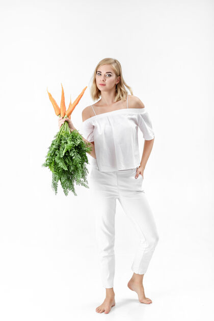一个一个穿着白衬衫的金发美女 手里拿着一根新鲜的胡萝卜 上面有绿叶背景.健康还有节食快乐年轻营养