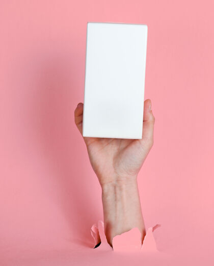 手臂女性手拿着白色盒子穿过撕破的粉色纸简约创意时尚理念手包装小