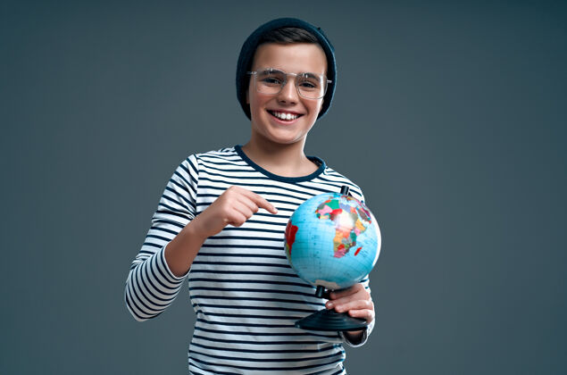 学习一个戴着帽子和眼镜的时髦聪明的小学生在一个灰色的地球上展示一个手指微笑小童年