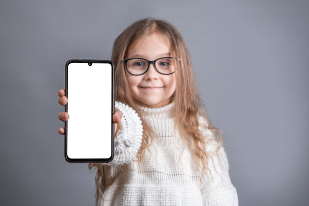 女性一个年轻漂亮的小女孩 金发飘逸的长发 穿着白色毛衣 在灰色的工作室里展示了一部白色空白屏幕的手机背景.地点为了文本.复制空间智能人技术