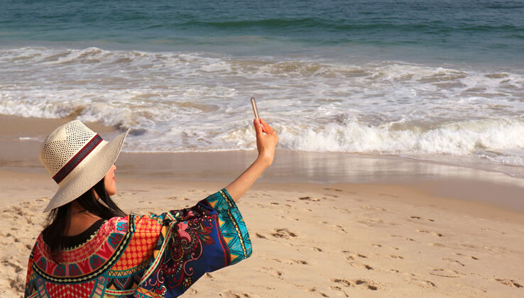 海浪撞击戴宽边帽的女人喜欢在波涛汹涌的海滩上自拍阳光水移动