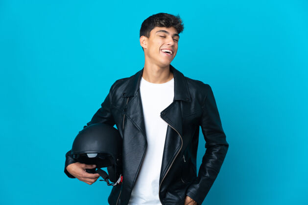 驾驶一个戴着摩托车头盔的年轻人在孤立的蓝色背景下大笑肖像摩托车手头盔