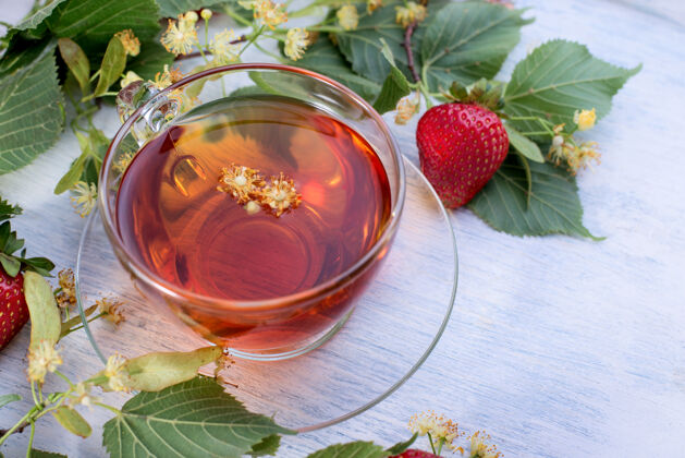 叶子一杯菩提茶 上面放着菩提花 树叶和草莓 放在一张旧的白色木制桌面上查看医疗保健热饮杯子替代品草药