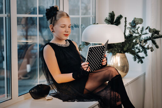 早晨一个穿着黑色连衣裙的时髦女孩坐在窗边的窗台上 手里拿着一本书复古窗台复古发型