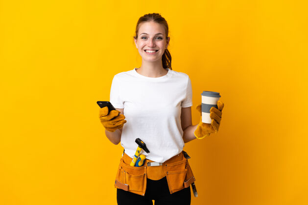 木工年轻的电工女孤立在黄色的背景下拿着咖啡带走和移动交谈电话电话