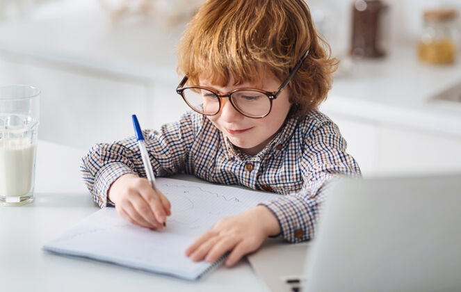大脑很少天才执着活泼的姜孩子戴着大眼镜 坐在白色的桌子旁 仔细地写下自己的想法休闲电脑书房