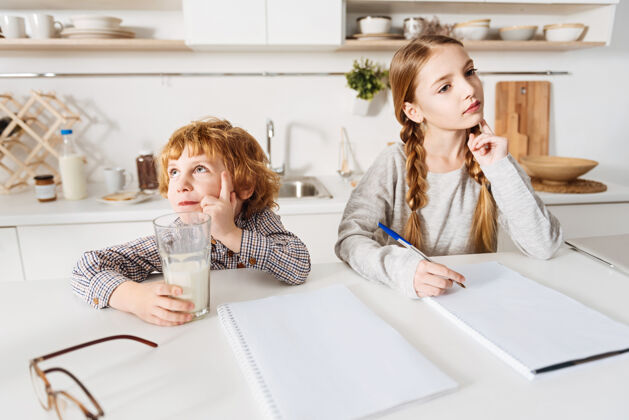 发展梦幻般的时间智慧有责任心的漂亮女孩在做家庭作业 她的弟弟在厨房里喝牛奶帮助学习教育