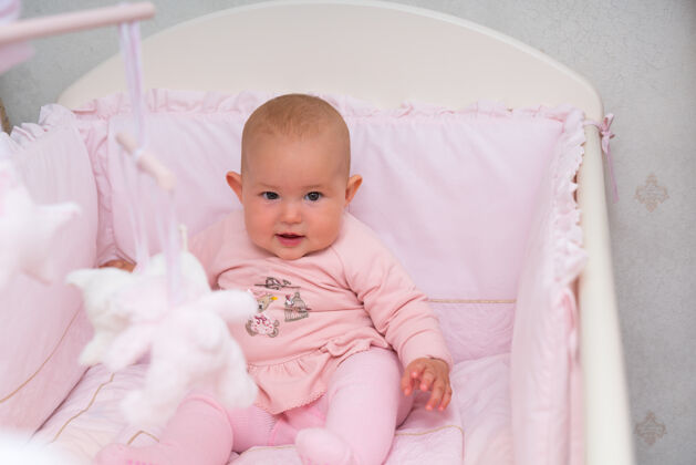 服装可爱的小女婴坐在粉红色的小床上 在近距离的镜头里好奇地看着镜头婴儿床看女孩