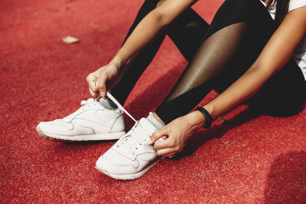 手特写镜头：一只雌性的腿坐在运动场的地上 在跑步前系鞋带女人体育场锻炼
