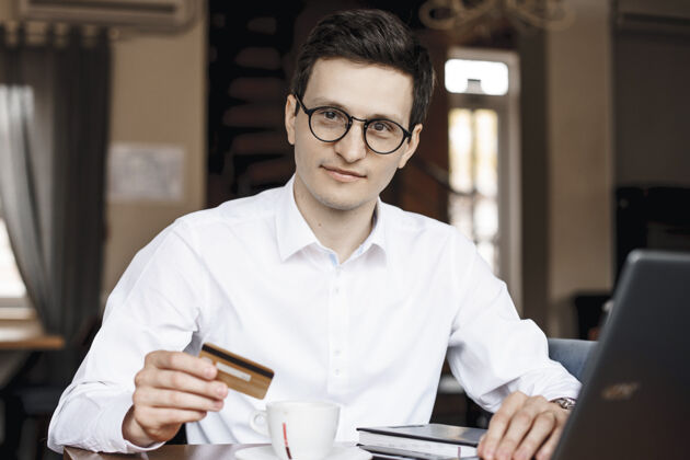 信贷年轻的商人坐在咖啡店里 手里拿着一张信用卡 看着相机消费主义自由职业者肖像