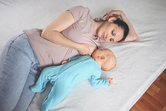 营养年轻的母亲躺在床上 抱着一个穿着蓝色连体衣的可爱的新生婴儿 用母乳给他喂奶女人母亲童年