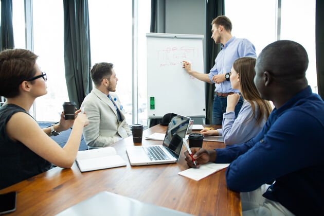 领导讨论公司进步自信年轻人站在白板旁边 指着图表 而他的同事坐在办公桌旁随意伙伴关系场所