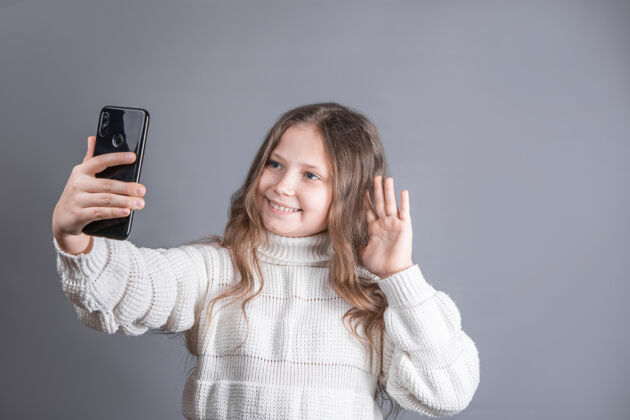 可爱一个年轻漂亮的小女孩的肖像 金发飘逸的长发 穿着白色毛衣 手持手机 在视频通讯中自拍自拍手机手机