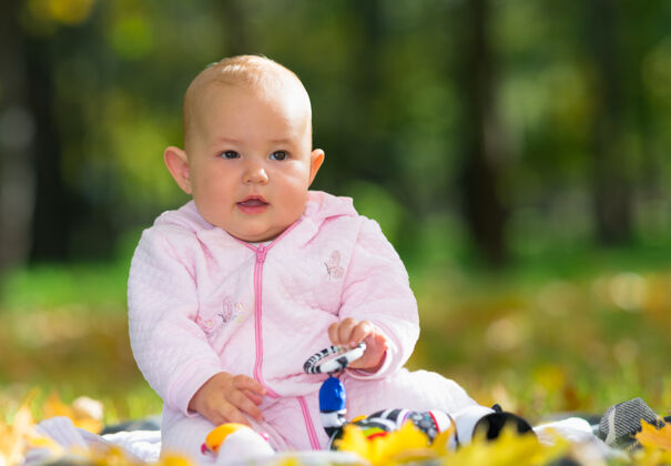 抬头可爱的粉红色女婴坐在草地上的地毯上 在公园里的黄色秋叶中玩玩具独自人女儿