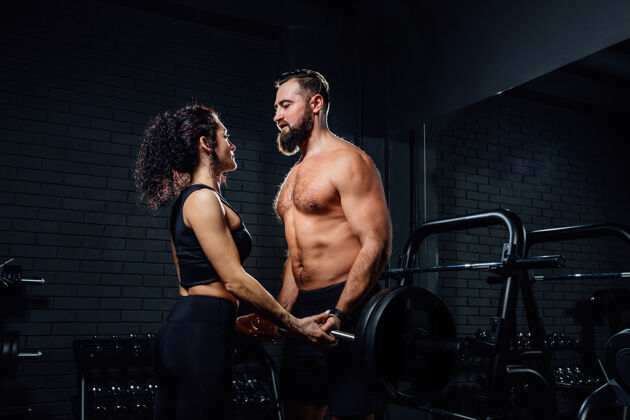 现代侧视图肌肉发达的胡须男子和美丽的女子看着对方 并提出沉重的杠铃一起在黑暗的健身房锻炼重量器械物理