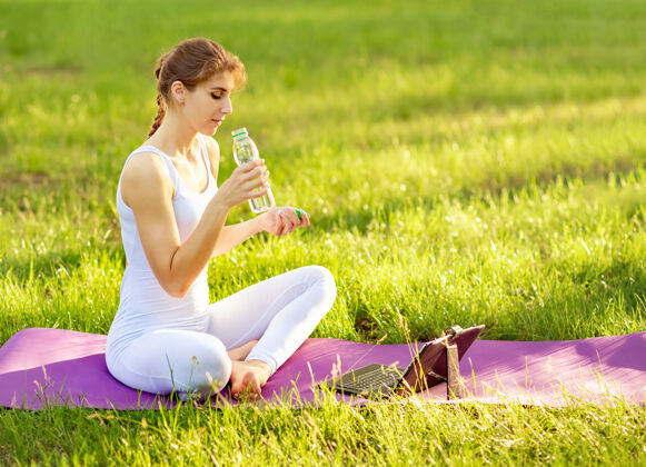 年轻人在公园的瑜伽垫上 一个微笑的女人拿着一瓶运动水瓶子普拉提女人