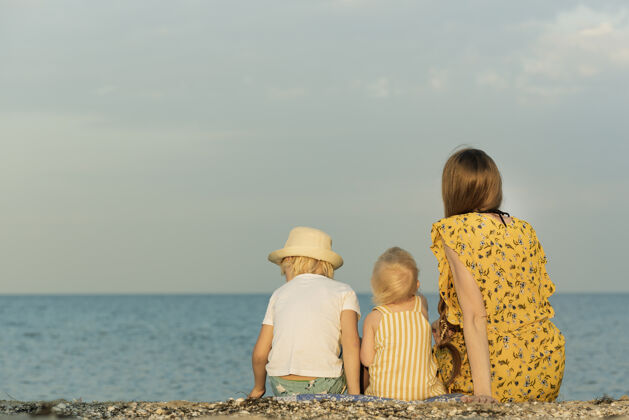 温柔母亲带着两个孩子坐在沙滩上望着窗外海背视图.summer带孩子度假假期海边沙滩