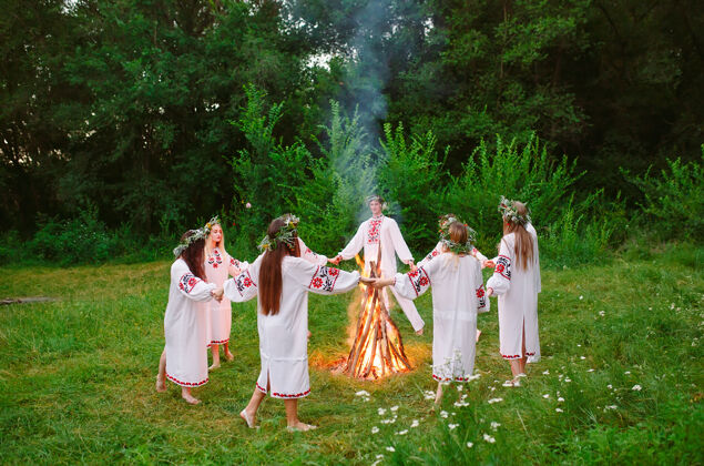 自然仲夏年轻穿着斯拉夫服装的人们在森林里围着篝火跳舞神圣乡村植物