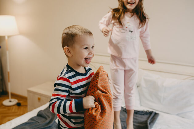 儿子可爱快乐的小孩抱着一个垫子在床上 而他的妹妹在早上跳朋友友谊孩子