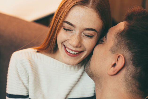 女人一位红发雀斑的可爱女性的特写照片 她闭上眼睛笑着 而她的男人正坐在沙发上亲吻她的脸颊微笑年轻浪漫