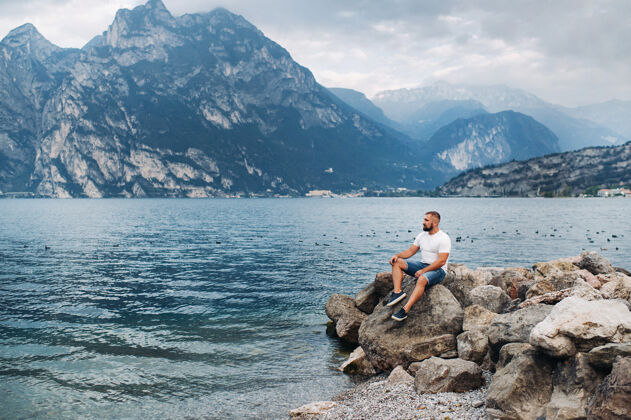 户外男性旅行者坐在阿尔卑斯山和湖泊背景下的岩石上嘉达假日酒店在意大利勇气年轻湖泊