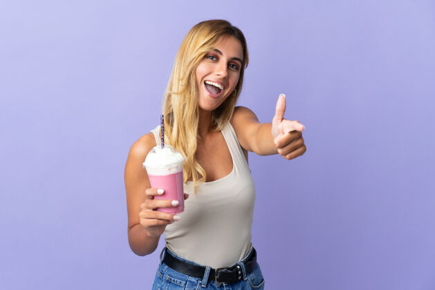 金发年轻的金发乌拉圭女人 拿着草莓奶昔 竖起大拇指站在紫色的地板上 因为发生了好事奶昔水果牛奶