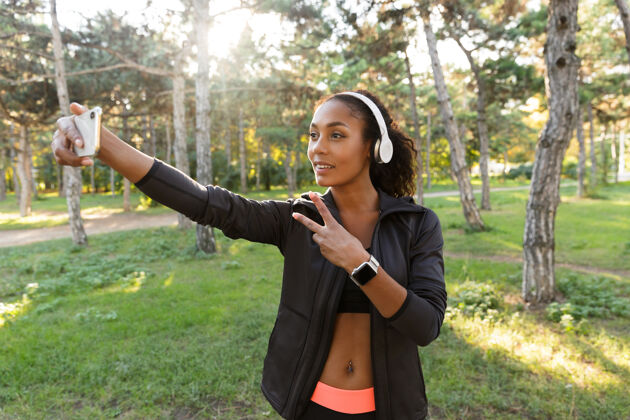 美国20多岁的快乐女人穿着黑色运动服 戴着耳机 在绿色公园散步时用手机自拍散步精力充沛运动服