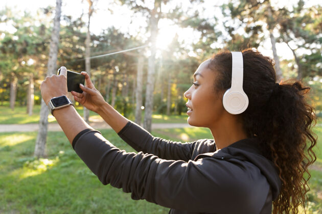 身体20多岁的美女穿着黑色运动服 戴着耳机 在绿色公园散步时用手机自拍运动锻炼精力充沛