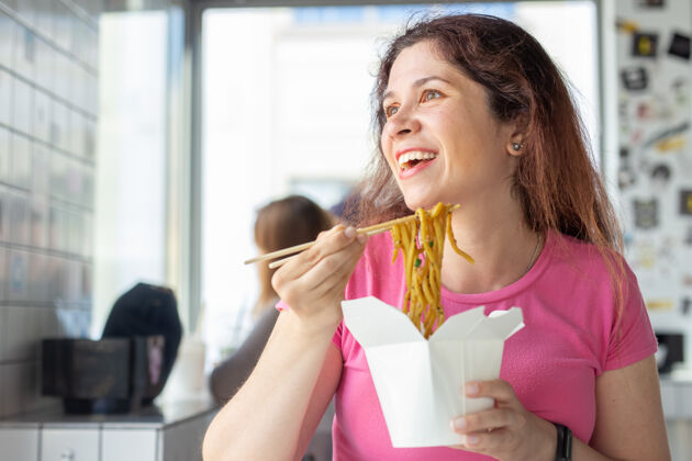 女人一个年轻开朗的女孩在咖啡馆吃中国面条 看着窗外的画像餐厅白种人咖啡馆