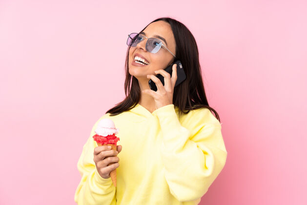 冰激凌年轻的黑发女孩拿着小号冰激凌在孤立的粉红色墙上拿着咖啡带走和手机甜点饥饿电话