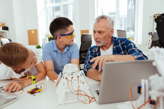 工程教育系统一位和蔼可亲的老教师一边向学生讲解机器人 一边和他们交谈老年人知识笔记本电脑