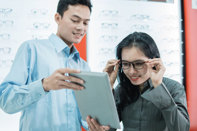 尝试一名店员站在眼镜店眼镜架展示柜的背景下 为试图戴眼镜观看平板电脑屏幕的女性顾客提供服务客户显示器微笑