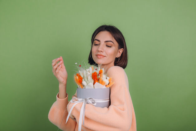 干花年轻女子穿着休闲桃色毛衣隔离在绿橄榄墙上手持橙白色花盒组成的棉花 吉普赛拉小麦和拉古鲁斯作为礼物开心惊喜时尚礼盒年轻